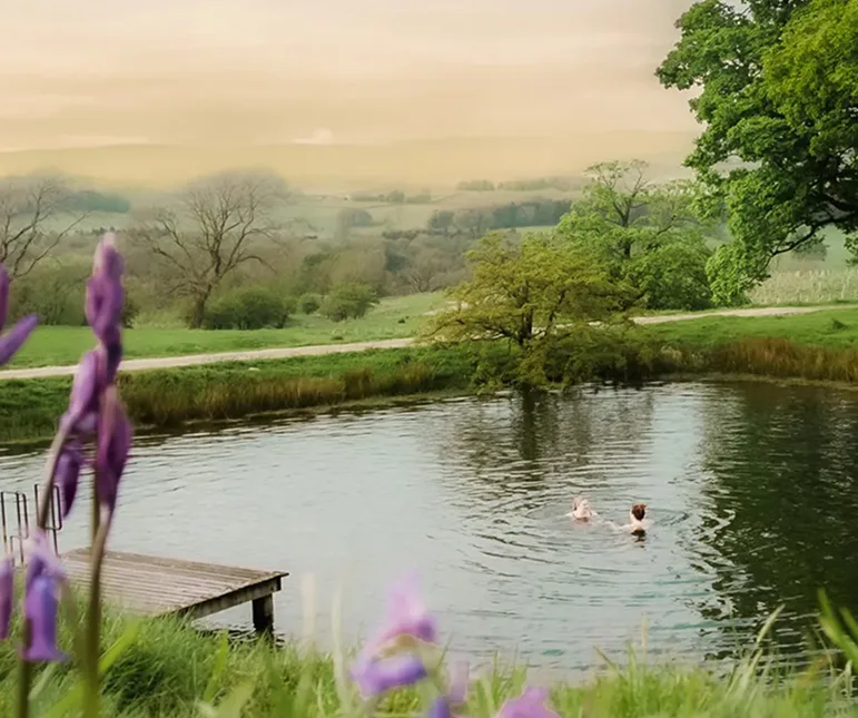 Two people wild swimming in a lake at dusk, bluebells in the foreground and the Yorkshire hills beyond
